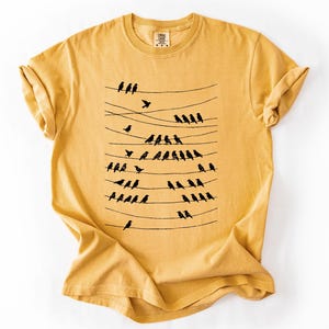 Op de afbeelding: Mosterdgeel T-shirt met een zwarte afbeelding van vogels op elektriciteitsdraden. Het ontwerp toont meerdere horizontale lijnen met vogels die zitten en vliegen. Het shirt heeft een ronde hals en korte mouwen.