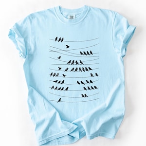 Op de afbeelding: Lichtblauw T-shirt met een zwarte afbeelding van vogels die op elektriciteitsdraden zitten. De vogels zijn in rijen gerangschikt, met enkele vogels die tussen de draden vliegen. Het shirt is gemaakt van zacht materiaal en heeft een ronde hals.