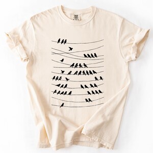 Op de afbeelding: Cr&egrave;mekleurig T-shirt met een zwarte afbeelding van vogels op elektriciteitsdraden. De vogels zijn in rijen gerangschikt, sommige vliegen. Het shirt heeft korte mouwen en een ronde hals.