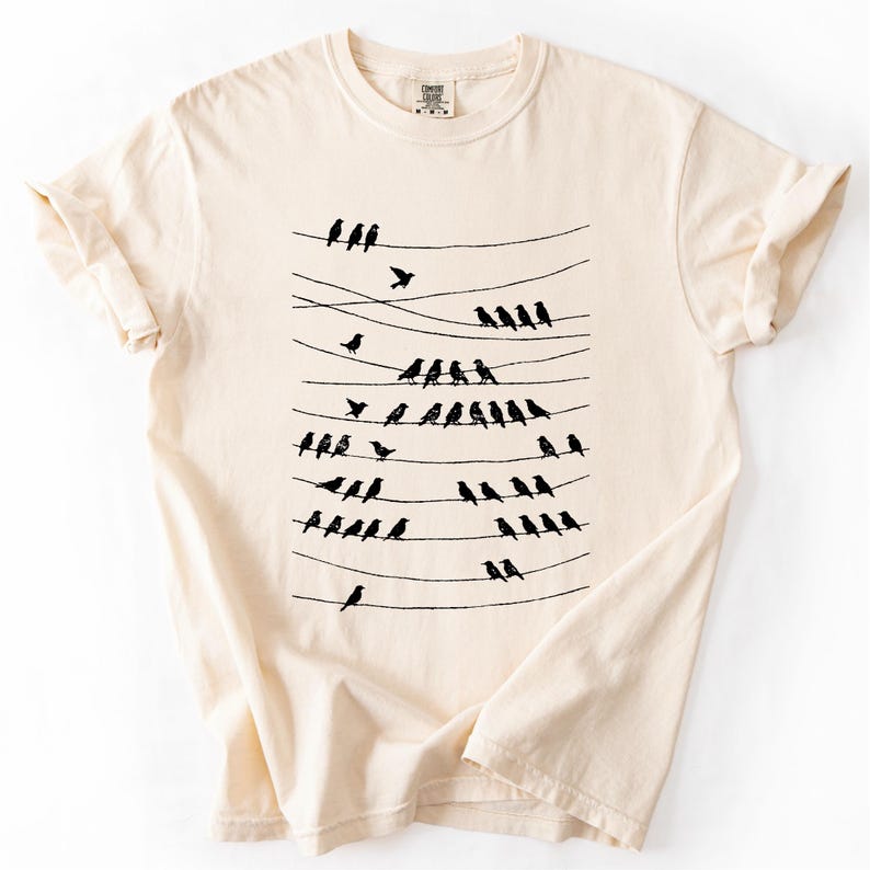 Op de afbeelding: Cr&egrave;mekleurig T-shirt met een zwarte afbeelding van vogels op elektriciteitsdraden. De vogels zijn in rijen gerangschikt, sommige vliegen. Het shirt heeft korte mouwen en een ronde hals.