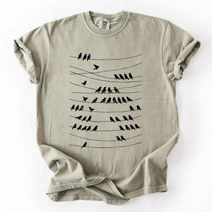 Op de afbeelding: Licht olijfgroen T-shirt met een zwarte afbeelding van vogels op elektriciteitsdraden. De vogels staan in rijen, sommige vliegen. Het shirt heeft een ronde hals en korte mouwen. Met Comfort Colors label.