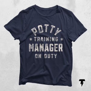 May include: Navy blue t-shirt with the text "POTTY TRAINING MANAGER ON DUTY" in distressed white lettering. The text is arranged in a stacked format with stars. A humorous and casual tee.