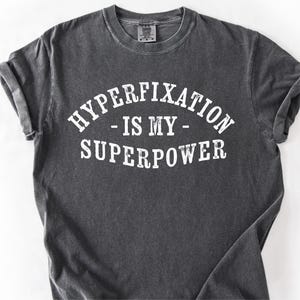 May include: Dark gray t-shirt with the text "HYPERFIXATION - IS MY - SUPERPOWER" in white, arched across the top and bottom. The shirt has a crew neck and short sleeves. The fabric appears soft and slightly faded.