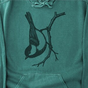 May include: Teal hoodie with a black and white illustration of a bird hanging upside down from a branch. The bird has a detailed wing pattern. The hoodie has a front pocket and a drawstring hood. The background is a solid teal colour.