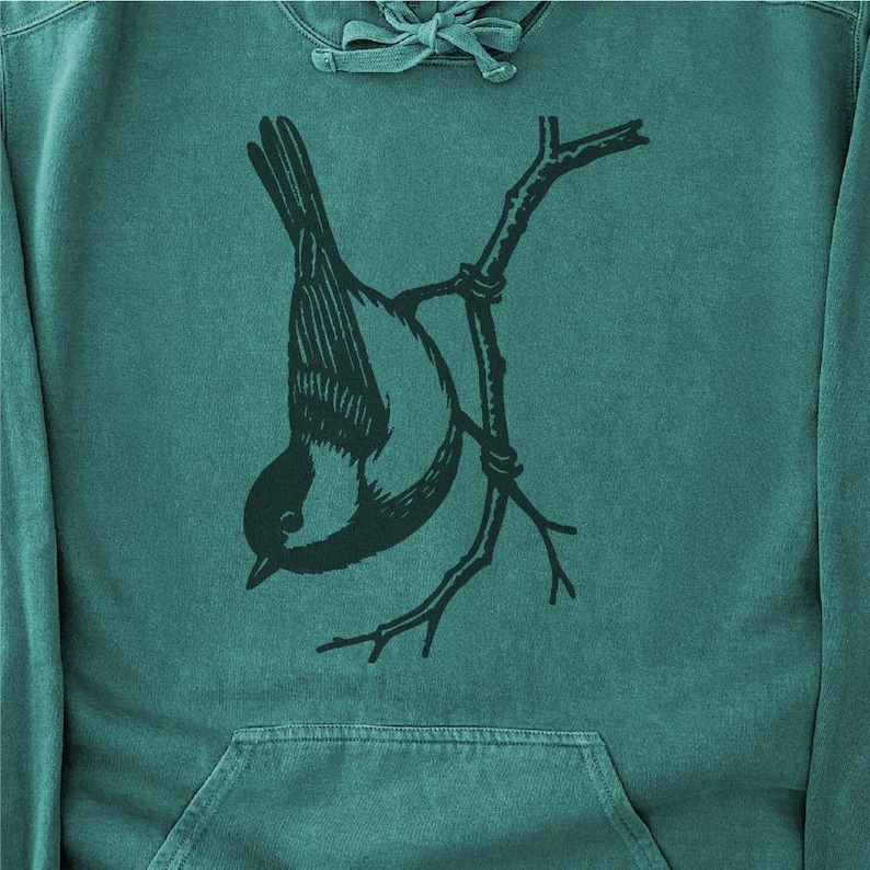 May include: Teal hoodie with a black and white illustration of a bird hanging upside down from a branch. The bird has a detailed wing pattern. The hoodie has a front pocket and a drawstring hood. The background is a solid teal colour.
