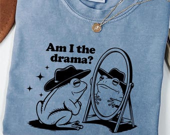 Op de afbeelding: Een blauw T-shirt met een zwart-witte illustratie van een kikker met een cowboyhoed die in een spiegel kijkt. De tekst "Am I the drama?" staat op het T-shirt gedrukt.