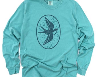 Könnte beinhalten: Türkisfarbenes Langarmshirt mit einem dunkelblauen Vogelmotiv in einem ovalen Rahmen. Der Vogel ist im Flug mit ausgebreiteten Flügeln dargestellt. Das Shirt besteht aus einem weichen Material und hat einen Rundhalsausschnitt.