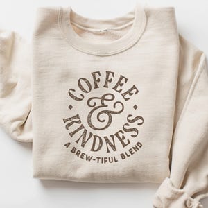 May include: A cream-coloured sweatshirt featuring the words "COFFEE & KINDNESS" in a brown, distressed font. The phrase "A BREW-TIFUL BLEND" is below. The sweatshirt is neatly folded.