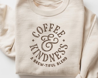 May include: A beige sweatshirt with the words "COFFEE & KINDNESS" in a brown, distressed font. Below the words is the phrase "A BREW-TIFUL BLEND". The sweatshirt is folded neatly.