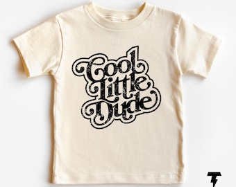 May include: Cream-colored toddler t-shirt with the phrase "Cool Little Dude" in a black, retro-style font. The text has a distressed effect, and the shirt has short sleeves and a crew neck.