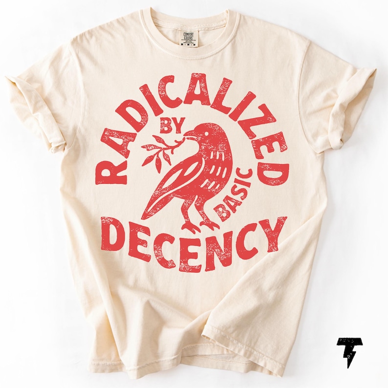 May include: Cream-colored t-shirt with rolled-up sleeves. The front features a distressed red graphic of a bird holding a sprig, encircled by the words "RADICALIZED BY BASIC DECENCY."