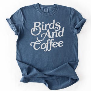 Könnte beinhalten: Ein blaues T-Shirt mit kurzen Ärmeln und Rundhalsausschnitt. Auf der Vorderseite befindet sich mittig ein weißer, im Used-Look gehaltener Schreibschriftzug mit dem Text "Birds And Coffee". Der Stoff hat ein leicht verwaschenes Aussehen.