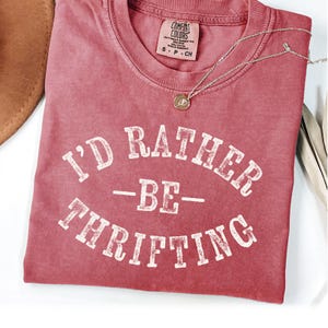 May include: A red t-shirt with the text "I'd Rather Be Thrifting" in white. The shirt is made of a soft, comfortable material and has a vintage look.