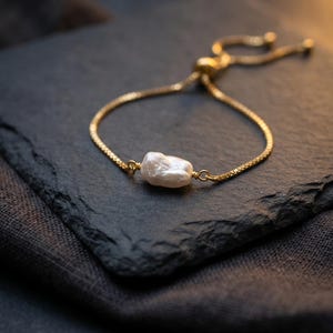 May include: A gold bracelet featuring a single, irregular white pearl. The bracelet sits on a dark grey, textured surface, against a dark backdrop. The chain is fine and adjustable, with a sliding clasp.