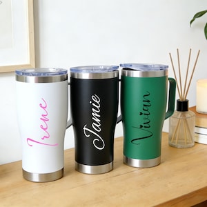 Puede incluir: Tres vasos personalizados en blanco, negro y verde, cada uno con asa y base plateada. El vaso blanco tiene el nombre "Irene" en escritura rosa, el vaso negro tiene "Jamie" en escritura blanca y el vaso verde tiene "Vivian" en escritura blanca.