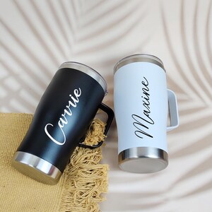 Puede incluir: Dos vasos personalizados, uno negro y otro blanco, cada uno con asa y base plateada. El vaso negro tiene el nombre "Carrie" en escritura blanca, mientras que el vaso blanco tiene "Maxine" en escritura negra. Los vasos est&aacute;n sobre una superficie beige.