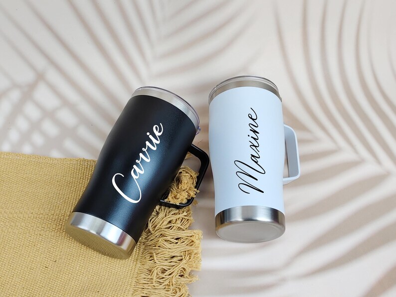 Puede incluir: Dos vasos personalizados, uno negro y otro blanco, cada uno con asa y base plateada. El vaso negro tiene el nombre "Carrie" en escritura blanca, mientras que el vaso blanco tiene "Maxine" en escritura negra. Los vasos est&aacute;n sobre una superficie beige.