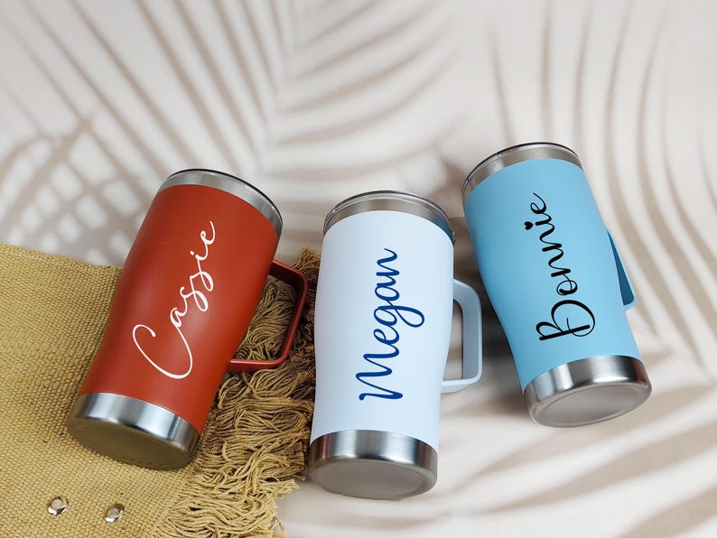 Puede incluir: Tres tazas de viaje personalizadas en rojo, blanco y azul. Cada vaso tiene un asa y una base y borde plateados. La taza roja est&aacute; inscrita con "Cassie", la taza blanca con "Megan" y la taza azul con "Bonnie".
