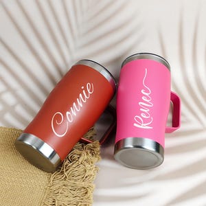 Puede incluir: Dos vasos personalizados, uno rojo y otro rosa, con detalles plateados. El vaso rojo tiene el nombre "Connie" en escritura blanca, mientras que el vaso rosa tiene "Renee". Ambos tienen asas y est&aacute;n sobre una esterilla tejida.
