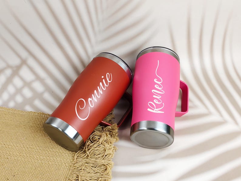 Puede incluir: Dos vasos personalizados, uno rojo y otro rosa, con detalles plateados. El vaso rojo tiene el nombre "Connie" en escritura blanca, mientras que el vaso rosa tiene "Renee". Ambos tienen asas y est&aacute;n sobre una esterilla tejida.