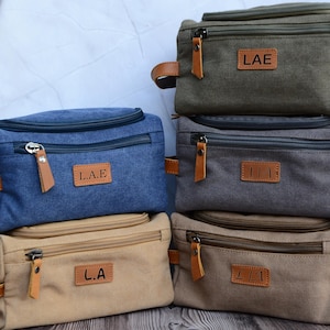 May include: Four canvas wash bags with leather accents and zipper closures. The bags are stacked on top of each other and have different initials on the front: LAE, J.J.A, L.A, and L/A.