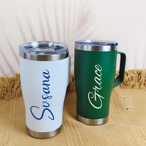 Puede incluir: Dos tazas de viaje de acero inoxidable con asas y tapas. Una es blanca con "Susana" en escritura azul, la otra es verde con "Grace" en escritura blanca. Ambas tienen borde y base plateados.