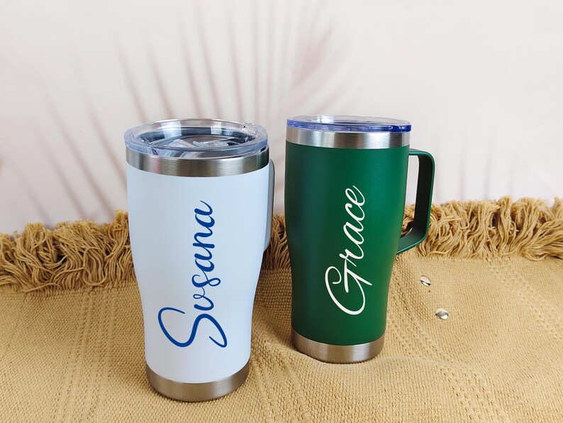 Puede incluir: Dos tazas de viaje de acero inoxidable con asas y tapas. Una es blanca con "Susana" en escritura azul, la otra es verde con "Grace" en escritura blanca. Ambas tienen borde y base plateados.