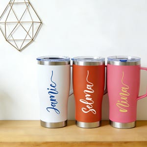 Puede incluir: Tres vasos personalizados con asas, en blanco, naranja-rojo y rosa. Cada vaso tiene un nombre en cursiva: Jamie (azul), Selma (blanco) y Nina (dorado). Los vasos son de acero inoxidable con tapas transparentes.