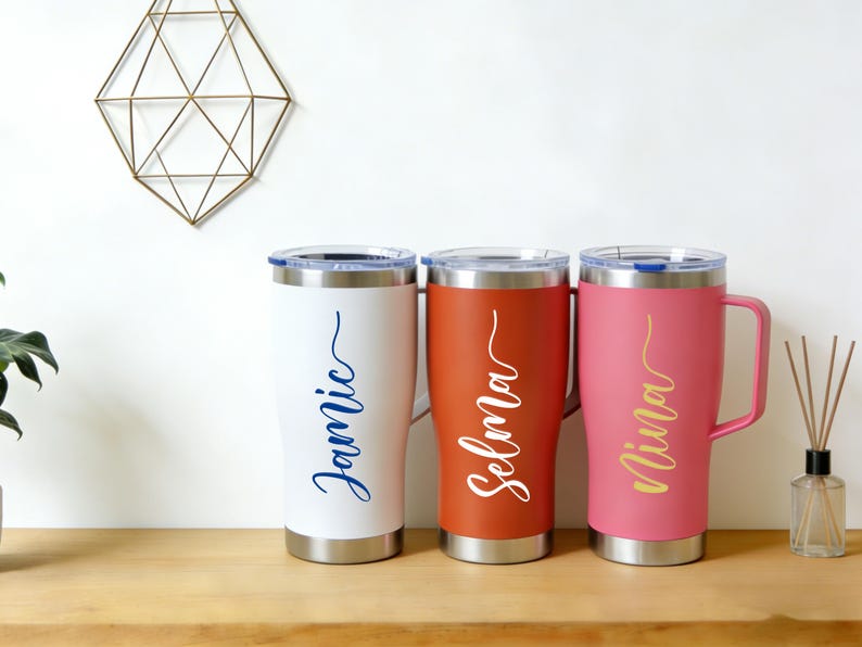 Puede incluir: Tres vasos personalizados con asas, en blanco, naranja-rojo y rosa. Cada vaso tiene un nombre en cursiva: Jamie (azul), Selma (blanco) y Nina (dorado). Los vasos son de acero inoxidable con tapas transparentes.
