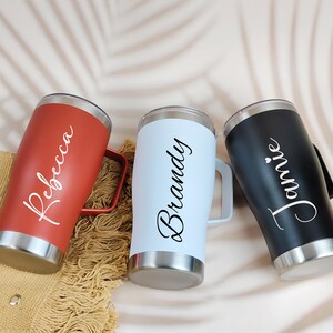 Puede incluir: Tres vasos aislados en rojo, azul claro y negro, cada uno con base y asa plateadas. Los nombres "Rebecca", "Brandy" y "Jamie" est&aacute;n escritos en cursiva en cada vaso. Los vasos est&aacute;n dise&ntilde;ados para mantener las bebidas fr&iacute;as o calientes.