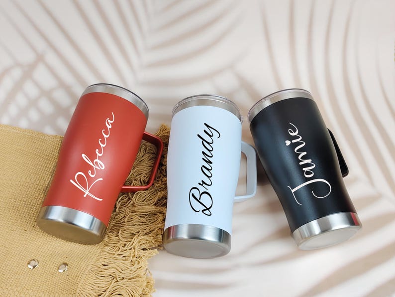 Puede incluir: Tres vasos aislados en rojo, azul claro y negro, cada uno con base y asa plateadas. Los nombres "Rebecca", "Brandy" y "Jamie" est&aacute;n escritos en cursiva en cada vaso. Los vasos est&aacute;n dise&ntilde;ados para mantener las bebidas fr&iacute;as o calientes.