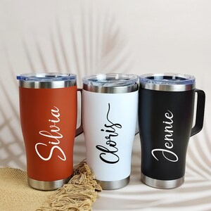 Puede incluir: Tres tazas de viaje personalizadas en &oacute;xido, blanco y negro. Cada vaso tiene un asa, tapa y base plateadas, con un nombre en escritura cursiva: "Silvia", "Cloris" y "Jennie". Las tazas est&aacute;n sobre una superficie neutra.