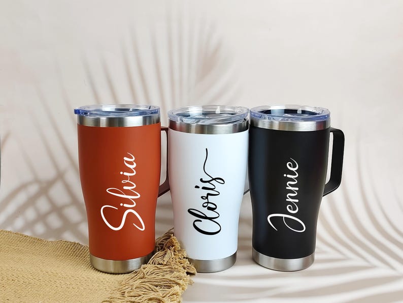 Puede incluir: Tres tazas de viaje personalizadas en &oacute;xido, blanco y negro. Cada vaso tiene un asa, tapa y base plateadas, con un nombre en escritura cursiva: "Silvia", "Cloris" y "Jennie". Las tazas est&aacute;n sobre una superficie neutra.