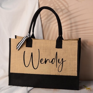 May include: A rectangular jute tote bag in natural tan with a black base. It has black handles and the name "Wendy" in black script. A striped ribbon is tied to one handle. The bag is suitable for shopping.