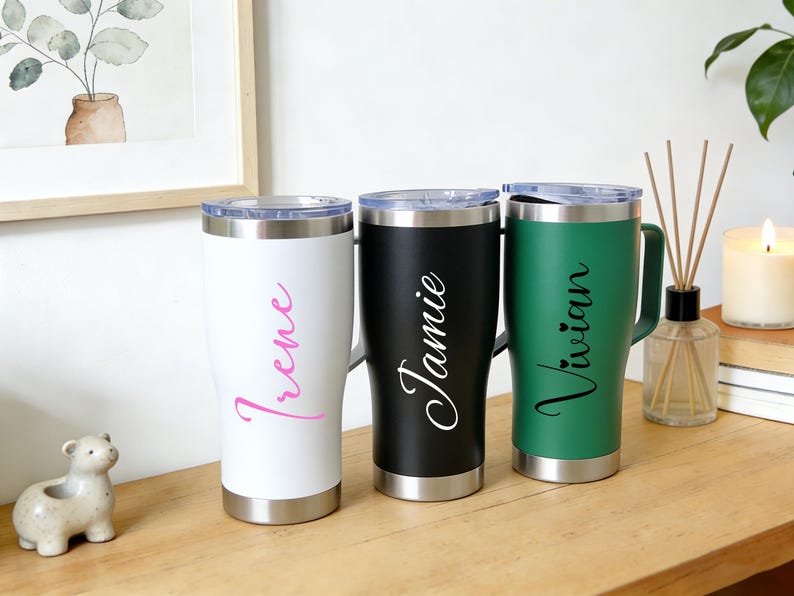 Puede incluir: Tres vasos personalizados en blanco, negro y verde, cada uno con asa y base plateada. El vaso blanco tiene el nombre "Irene" en escritura rosa, el vaso negro tiene "Jamie" en escritura blanca y el vaso verde tiene "Vivian" en escritura blanca.