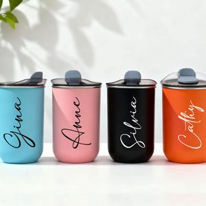 May include: Four insulated tumblers in blue, pink, black, and orange, each with a name in a cursive font. The tumblers have a gray lid and are designed to keep beverages hot or cold. The names are Gina, Anne, Silvia, and Cathy.