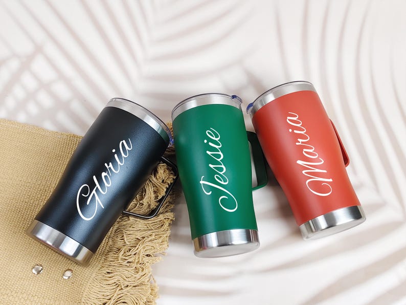 Puede incluir: Tres vasos personalizados de acero inoxidable en negro, verde y rojo. Cada vaso tiene un asa y un borde plateado. Los nombres "Gloria", "Jessie" y "Maria" est&aacute;n escritos en blanco en cada vaso.