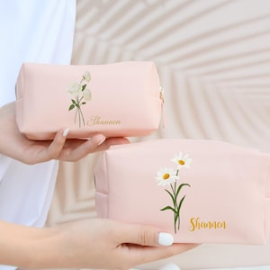 May include: Two pink zippered cosmetic bags with floral designs. The top bag has a white flower design and the bottom bag has a white daisy design. Both bags have the name "Shannon" printed in gold lettering.