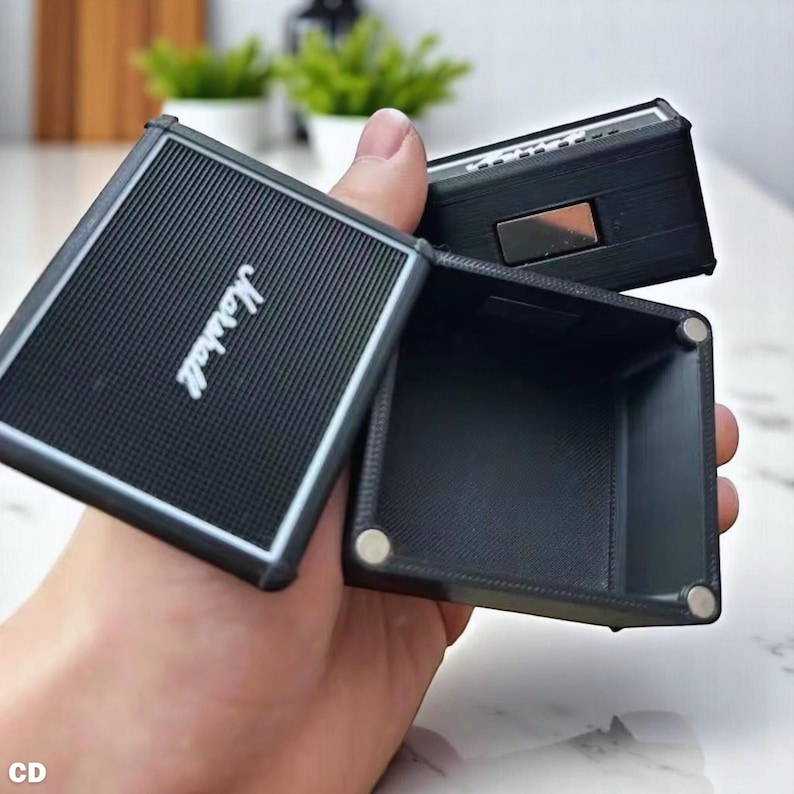 以下が含まれることがあります： 白のMarshallロゴが入った黒い箱型の容器。容器は開いており、黒い内部が見えます。容器はテクスチャ加工された表面とシルバーのトリムが付いています。容器は手で持たれています。