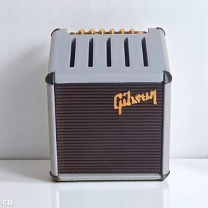 以下が含まれることがあります： 金色の「Gibson」の文字が入った、グレーと茶色のミニチュアアンプ。上部には縦のスロットと金色のノブがあります。前面には、濃い茶色のテクスチャードスピーカーエリアがあります。