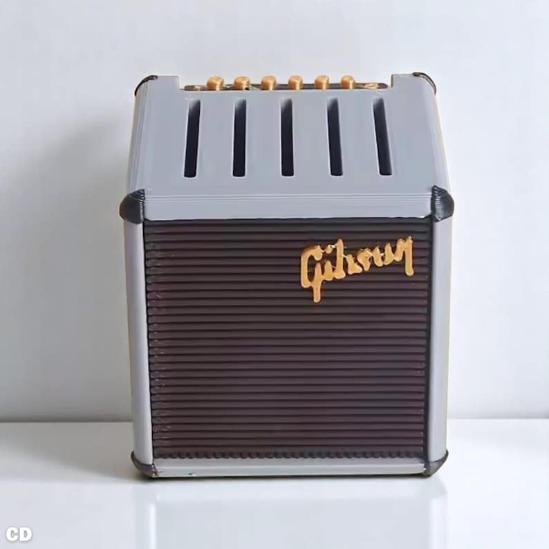 以下が含まれることがあります： 金色の「Gibson」の文字が入った、グレーと茶色のミニチュアアンプ。上部には縦のスロットと金色のノブがあります。前面には、濃い茶色のテクスチャードスピーカーエリアがあります。
