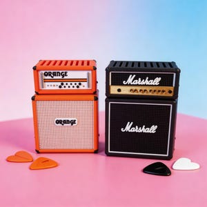 May include: Two miniature guitar amplifier replicas, one orange with "ORANGE" text and the other black with "Marshall" text. Several heart-shaped and guitar pick-shaped accessories are scattered in front of the amps. The background is a gradient of pink and blue.