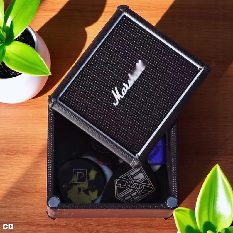 以下が含まれることがあります： 白い「Marshall」のロゴが付いた、スピーカーのような蓋が付いた黒と茶色の箱。箱は開いており、中には様々なアイテムが入っています。箱は木製の表面に置かれ、背景には植物があります。