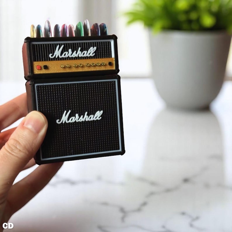 以下が含まれることがあります： 黒と金のMarshallアンプ型ペン立て。白い文字で「Marshall」と書かれています。ペン立てには、さまざまな色のペンがいくつか入っています。ペン立ては人が持っています。