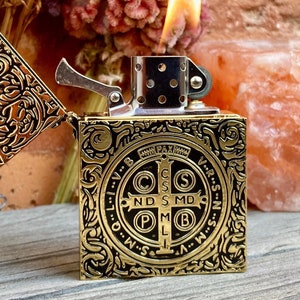 May include: A gold-colored lighter with a black and gold design. The lighter features a religious symbol with the words "V.R.S.N.S.M.D.O.L.M.B.V.R.S.N." around the outside. The lighter is open and the flame is visible.