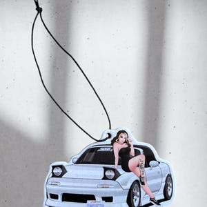 May include: Air freshener depicting a light blue car with a woman in a black dress. The car has the text "FEMKEYSETTEN" on the front. A black string is attached for hanging. The background is a light grey.