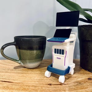 May include: A small white medical device with blue and purple detailing, including a screen and the word "Vivid". A dark green and black mug is positioned to the left.