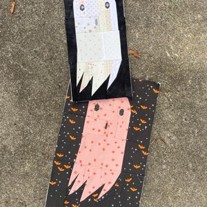 May include: Two handmade Halloween ghost-shaped fabric decorations. One ghost is white with a black background, and the other is pink with a black background decorated with orange bats and stars. The ghosts have button eyes and jagged bottoms.