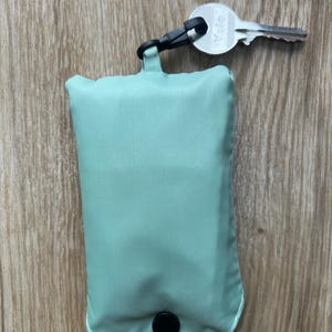 May include: A folded, pale green reusable shopping bag with a black press stud closure. A black clip and a Yale key are attached to the bag. The bag is made of a smooth, lightweight material.