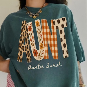 May include: A dark teal short-sleeve t-shirt featuring the word AUNT in large letters with varied patterns including leopard print, floral, orange gingham, and brown polka dots. Below the word AUNT, the text Auntie Sarah is printed in white.